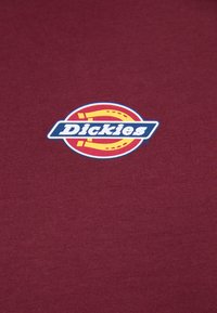 Dickies logo sa žutim potkovićom i bijelim tekstom na tamnocrvenoj tkanini.