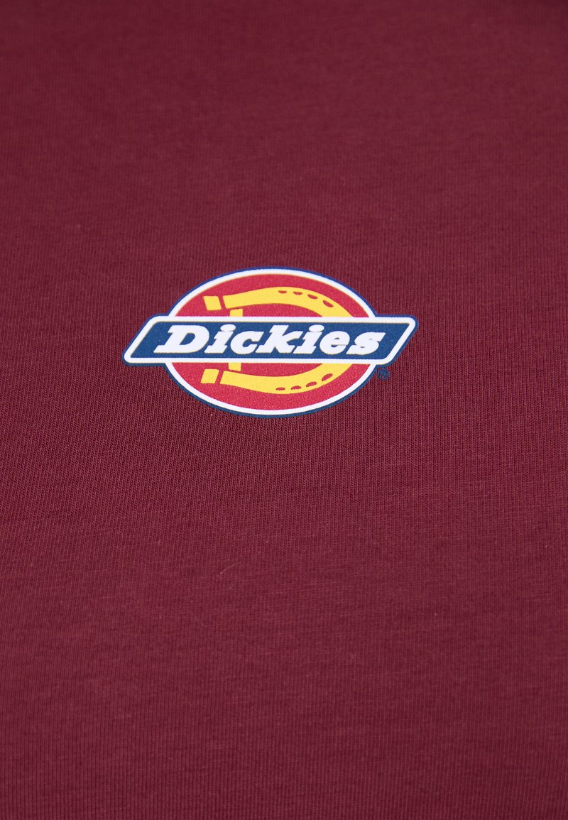 Logo Dickies avec un fer à cheval jaune et du texte blanc sur un fond en tissu bordeaux.