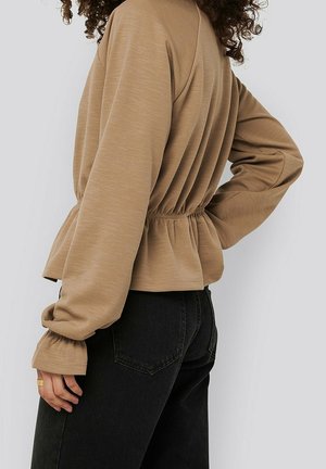 Kvinde iført beige bluse med indsnøring ved taljen og lange ærmer samt sorte jeans, stående med den ene hånd på hoften mod en ensfarvet, lys baggrund.