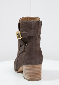 Bottine en suède marron avec un talon empilé, dotée de deux boucles dorées réglables et d'une fermeture éclair latérale pour un enfilage facile.