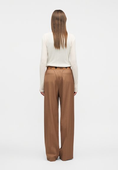 Femme debout, vue de dos, portant un haut blanc à manches longues et un pantalon large marron avec une ceinture de marque.