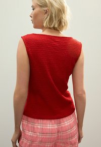 Débardeur rouge en tricot sans manches avec une finition texturée, présentant un large décolleté et une coupe décontractée, porté avec un bas à carreaux rose.