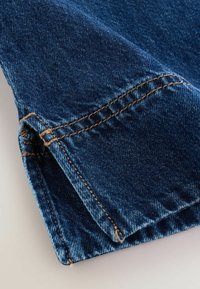 Prim-plan al tivului unei perechi de blugi din denim albastru, cu cusături portocalii vizibile și marginea țesăturii pliată pe un fundal deschis la culoare.