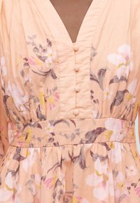 Ροδακινί λουλουδένιο φόρεμα με V-ντεκολτέ, κουμπιά καλυμμένα με ύφασμα μπροστά, σφηκοφωλιά στη μέση και πολύχρωμα λουλούδια σε ροζ, λευκούς και γκρι τόνους.