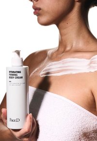 Bottiglia cilindrica bianca di crema idratante per il corpo con erogatore, etichettata "Crema Idratante e Rassodante per il Corpo", tenuta contro la pelle nuda con la crema applicata.