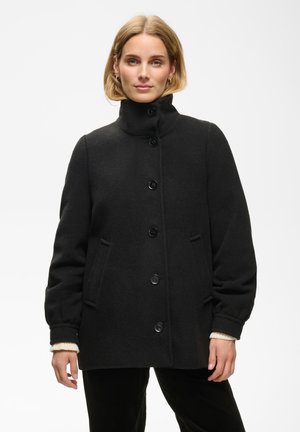 Femme portant un manteau en laine noir boutonné avec col montant, mains détendues le long du corps, sur un fond clair uni.