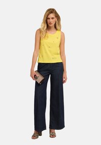 Top giallo senza maniche con decorazioni floreali, abbinato a pantaloni in denim scuri a gamba larga. Gli accessori includono una pochette beige e sandali con motivo.