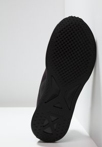 Suela de zapatilla deportiva negra, con una superficie de goma texturizada que presenta patrones circulares y hexagonales. Logo embosado en el área del talón.