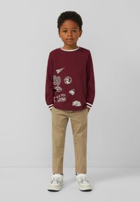 Bordeaux lange mouwen T-shirt met witte accenten en verschillende bedrukte patches; gecombineerd met beige broek en witte sneakers. Minimalistisch ontwerp.