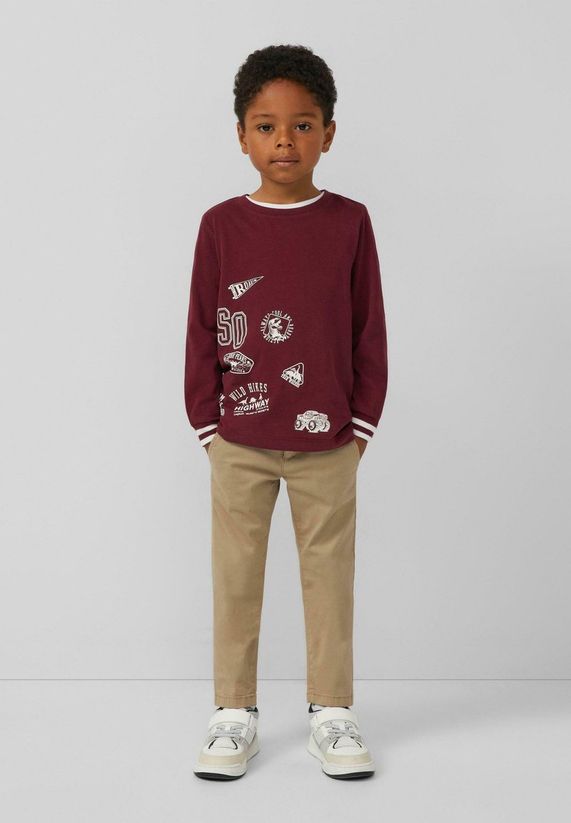 Bordeaux lange mouwen T-shirt met witte accenten en verschillende bedrukte patches; gecombineerd met beige broek en witte sneakers. Minimalistisch ontwerp.