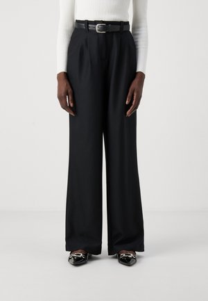 Pantalon noir à jambes larges en tissu lisse, associé à une fine ceinture noire avec une boucle en métal argenté, porté avec des mocassins noirs brillants.