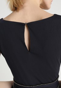 Schwarzes Kleid mit einem V-förmigen Ausschnitt am Rücken, ausgestattet mit einem kleinen Knopfverschluss und strukturiertem Stoff. Perlenakzent an der Taille.