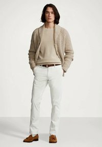 Jovem de pé com as mãos nos bolsos, vestindo um suéter de malha bege, um cardigan, calças brancas, cinto castanho e mocassins castanhos com franja.