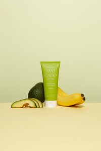 Maschera nutriente per il cuoio capelluto in un tubo verde brillante con tappo bianco, circondata da un avocado, una banana e fette di avocado su una superficie gialla.
