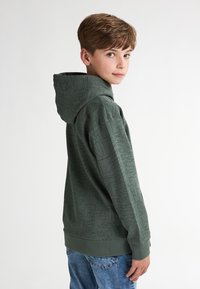 Zelená mikina s texturovaným vzorem, klokání kapsou a nastavitelnou kapucí; v kombinaci s modrými džíny.