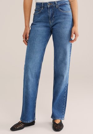 Naine, kes kannab keskmise vöökohaga sirge lõikega siniseid teksapükse ja musti punutud tasaseid kingi, seisab ühevärvilise beeži tausta ees.