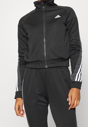 Personne portant un survêtement Adidas noir avec une veste zippée et un pantalon assorti avec des bandes blanches sur les manches et les jambes.