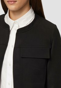 Schwarze Jacke mit Rundhalsausschnitt, mit einer Brusttasche und einer glatten Textur, über einem weißen Hemd mit Kragen getragen.