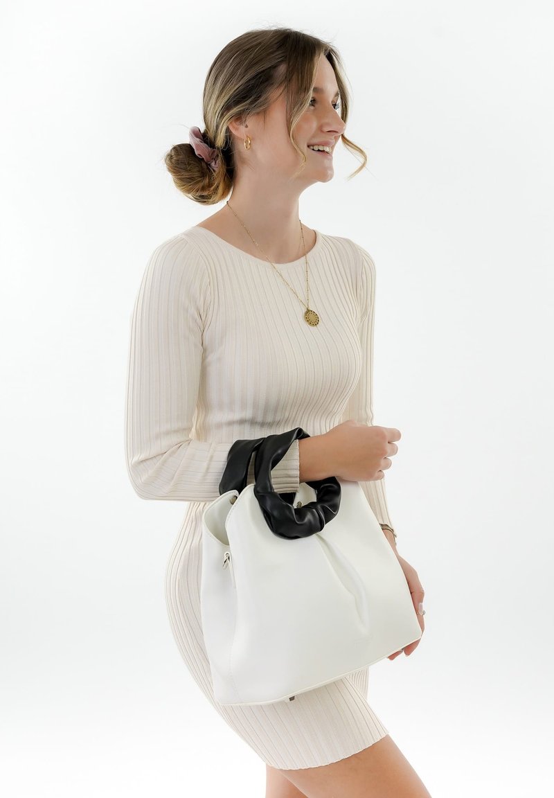 Lächelnde Frau in einem cremefarbenen, gerippten Kleid hält eine weiße Handtasche mit schwarzen Griffen, trägt goldenen Schmuck und ein pinkfarbenes Haargummi.