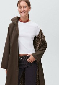 Joven mujer sonriendo, vestida con una blusa corta blanca, jeans oscuros con tachuelas y un abrigo oscuro con forro de leopardo, con una mano apoyada en la cadera.