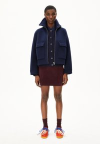 Chaqueta de lana azul marino con un cuello alto, dos bolsillos frontales, combinada con una falda de pana burdeos y zapatillas deportivas naranjas con acentos azules.