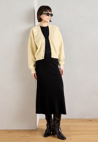 Cardigan en tricot crème sur une robe noire, texture côtelée, silhouette ajustée, associé à des bottes noires hauteur genou et des lunettes de soleil angulaires.