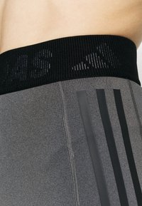 Šedé sportovní šortky mají texturovaný černý pas s reflexním nápisem, vertikální černé pruhy po stranách a viditelné švy.