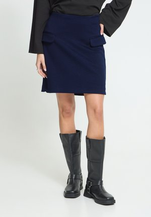 Femme portant une jupe bleu marine avec des poches à rabat à l'avant et des bottes noires montantes jusqu'aux genoux, debout avec une main sur la hanche.