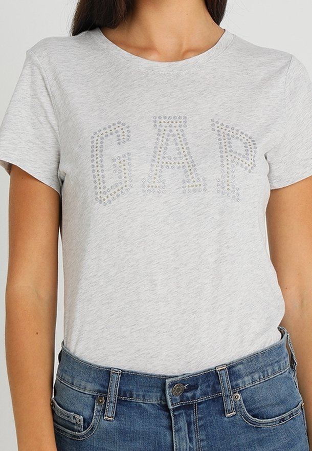 Femme portant un t-shirt gris clair avec le mot "GAP" écrit en petits clous argentés, associé à un jean en denim bleu.