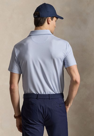 Άνδρας με ανοιχτό μπλε κοντομάνικο πουκάμισο πόλο με υφή, navy ζώνη, navy παντελόνι και navy καπέλο baseball, στραμμένος από πίσω.