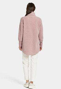 Fleece-Pullover in sanftem Hellrosa mit hohem Kragen und langen Ärmeln, mit abgerundetem Saum und gerippten Ärmelbündchen. Getragen mit weißen Hosen.