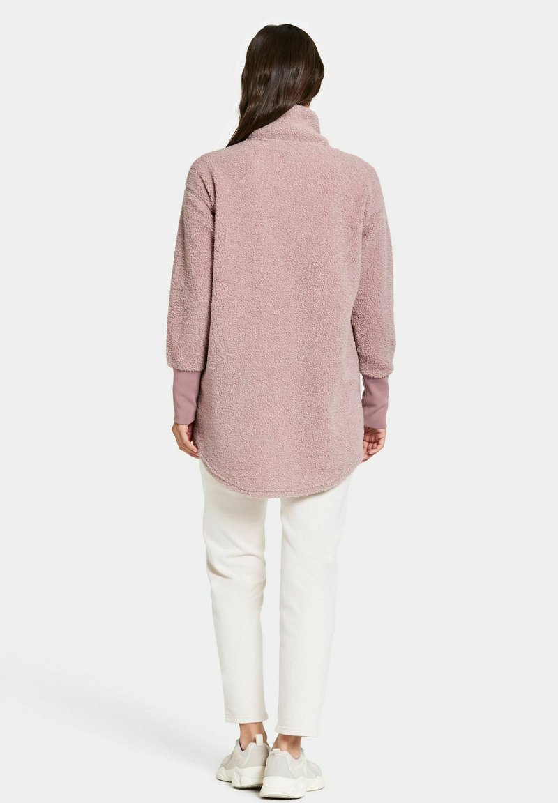 Fleece-Pullover in sanftem Hellrosa mit hohem Kragen und langen Ärmeln, mit abgerundetem Saum und gerippten Ärmelbündchen. Getragen mit weißen Hosen.