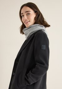 Cappotto nero con un design strutturato, caratterizzato da un ampio colletto, chiusura con bottoni e una toppa con logo sulla manica. Accompagnato da una sciarpa grigia.