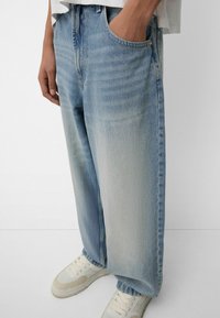 Hellblaue Denim-Jeans im entspannten Schnitt, mit hoher Taille, subtilen Verblasungen und einem klassischen Fünf-Taschen-Design. Getragen mit weißen Sneakers.