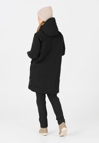 Svart lång parka i slitstarkt material, med huva, sidofickor och en minimalistisk design, kombinerat med svarta byxor och beige stövlar.