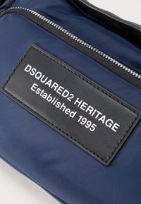 Bolso azul marino con parche de cuero negro que muestra texto blanco "DSQUARED2 HERITAGE Established 1995" debajo de una cremallera plateada.