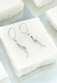 Silverörhängen med bladsform, med en böjd design, glittrande detaljer och en klämfäste, uppställda på marmorytor.