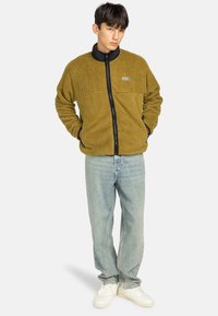 Fleecejack in olijfgroen met een zwarte kraag en rits aan de voorkant. Heeft een logo-opdruk, gestructureerde stof en is gecombineerd met lichtkleurige denim jeans.