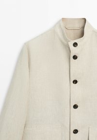Beige linen jacket with stand collar and five dark buttons down the front, showing upper torso and left sleeve.