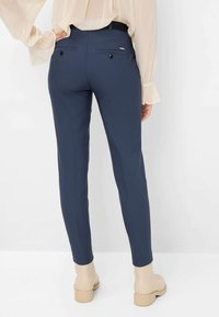 Pantalons ajustés bleu marine, coupe slim, dotés de deux poches arrières avec des boutons noirs, portés avec des bottines beiges et un haut léger et transparent.