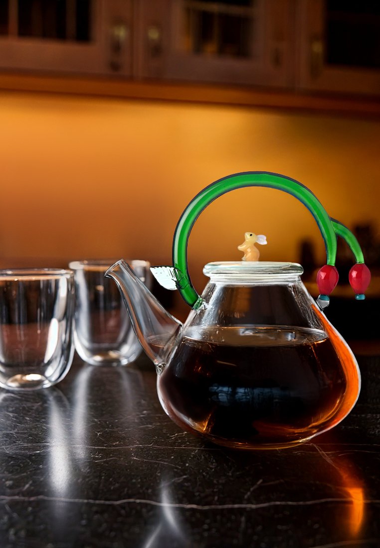 Szklany czajnik z zakrzywioną zieloną rączką, ozdobiony małym żółtym zajączkiem. Zawiera ciemną herbatę, obok znajdują się trzy przezroczyste filiżanki.