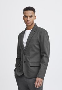 Dunkelgrauer Blazer mit glatter Textur, Ein-Knopf-Verschluss und zwei Fronttaschen. Über einem weißen T-Shirt getragen, schlank geschnitten.