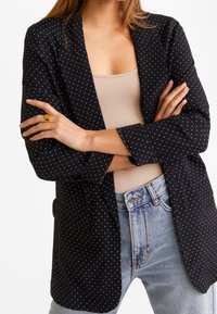 Schwarzer Blazer mit weißen Punkten, leichtes Material, offenes Vorderteil, Dreiviertelärmel, kombiniert mit einem beigen Top und blauen Jeans.