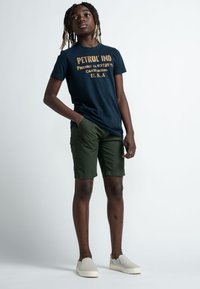 T-shirt blu marina con testo giallo, pantaloni corti verde oliva e scarpe slip-on grigie. Modello in piedi con le mani in tasca, sfondo neutro.