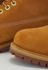 Tan kleurige suède laarzen met gouden oogjes, brown veters en een robuuste rubberen zool met diepe noppen; stiksel detail aan de bovenkant.