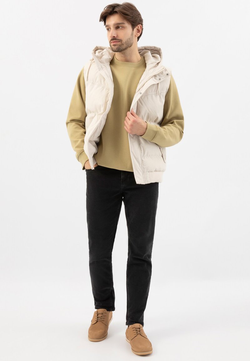 Mann trägt beige Kapuzen-Steppweste, hellbraunen Pullover, schwarze Jeans und beige Schuhe, steht mit einer Hand in der Tasche und schaut seitlich.