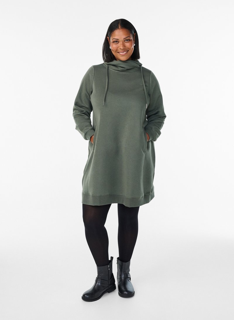 Smiling woman standing with hands in pockets wearing a green long-sleeve hooded dress, black tights, and black ankle boots.