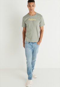 Camiseta corta de manga verde claro con un logo amarillo en el pecho, combinada con jeans azul claro y zapatillas blancas. Textura suave.