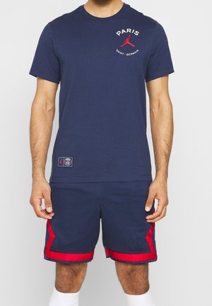 Homme portant un t-shirt bleu marine Paris Saint-Germain avec un logo Jordan rouge et un short bleu marine assorti avec bordure rouge.