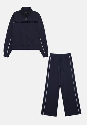 Survêtement bleu marine avec veste zippée et pantalons à jambes larges. Présente un liseré blanc le long de la poitrine et des pantalons pour un contraste. Confectionné dans un tissu doux.